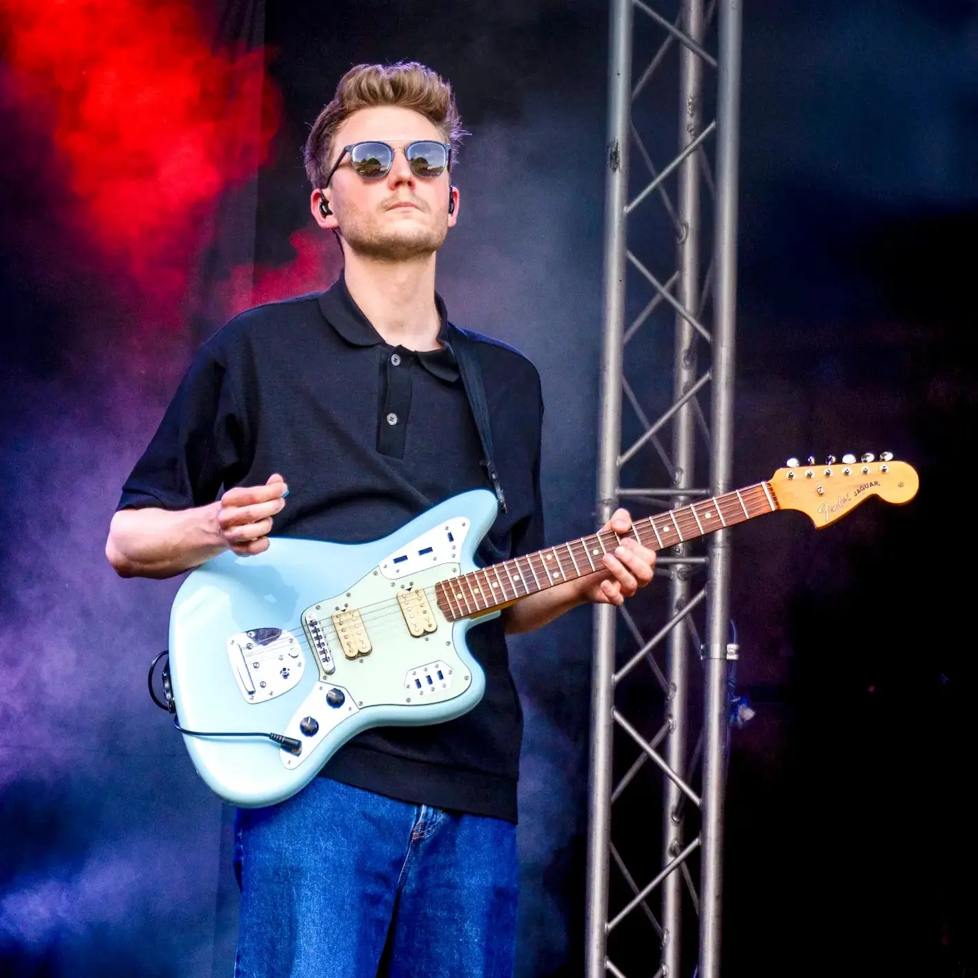 Gitarrist mit Sonnenbrille spielt hellblaue E-Gitarre auf einer Bühne im bunten Licht.