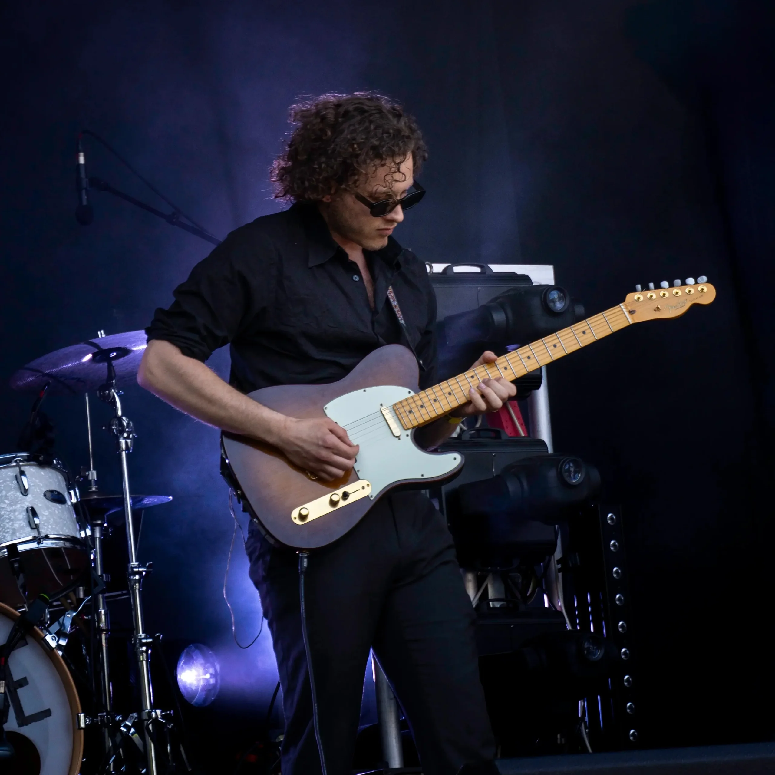 Gitarrist mit Sonnenbrille spielt E-Gitarre auf einer Bühne mit Schlagzeug im Hintergrund.
