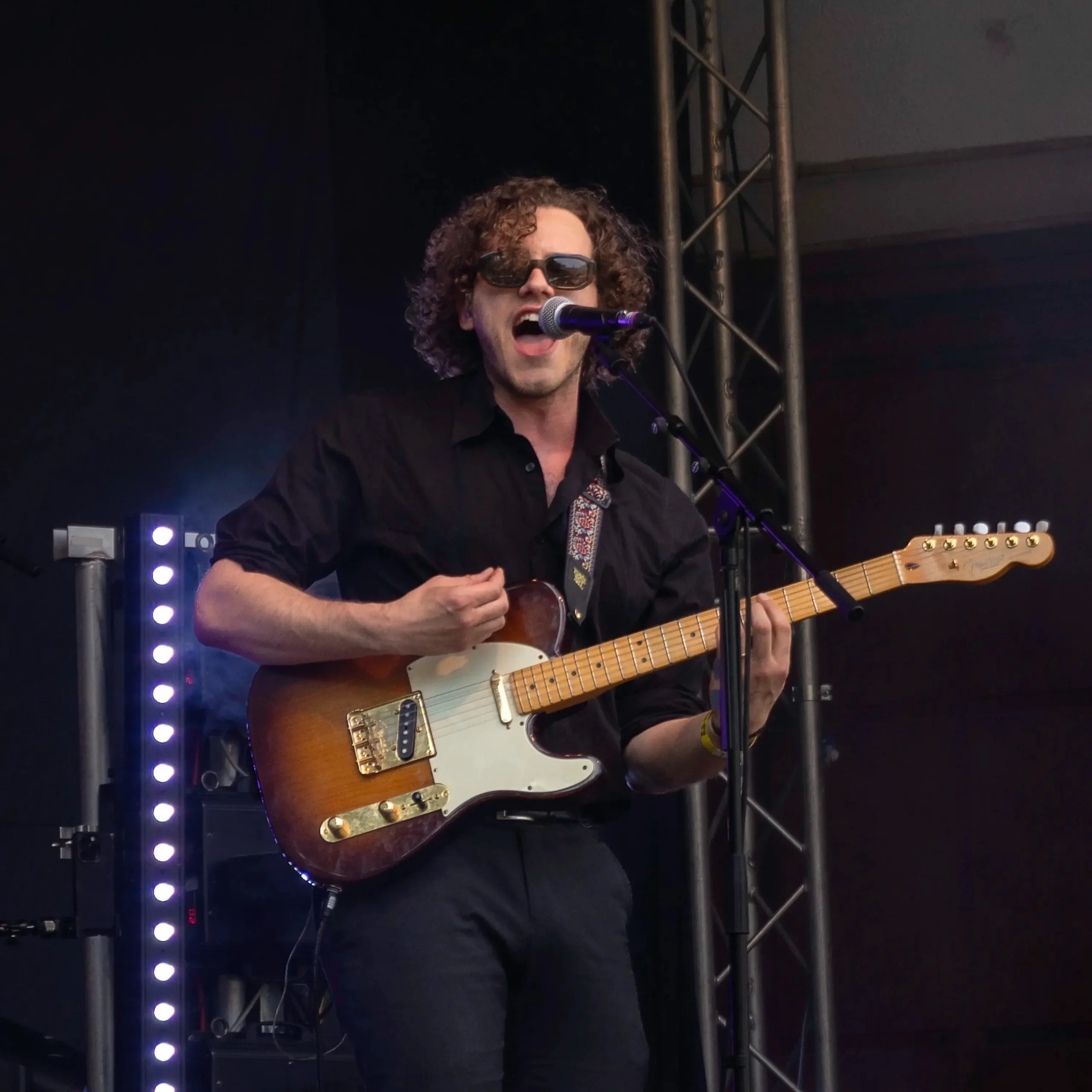 Gitarrist mit Sonnenbrille singt und spielt E-Gitarre auf einer Bühne.