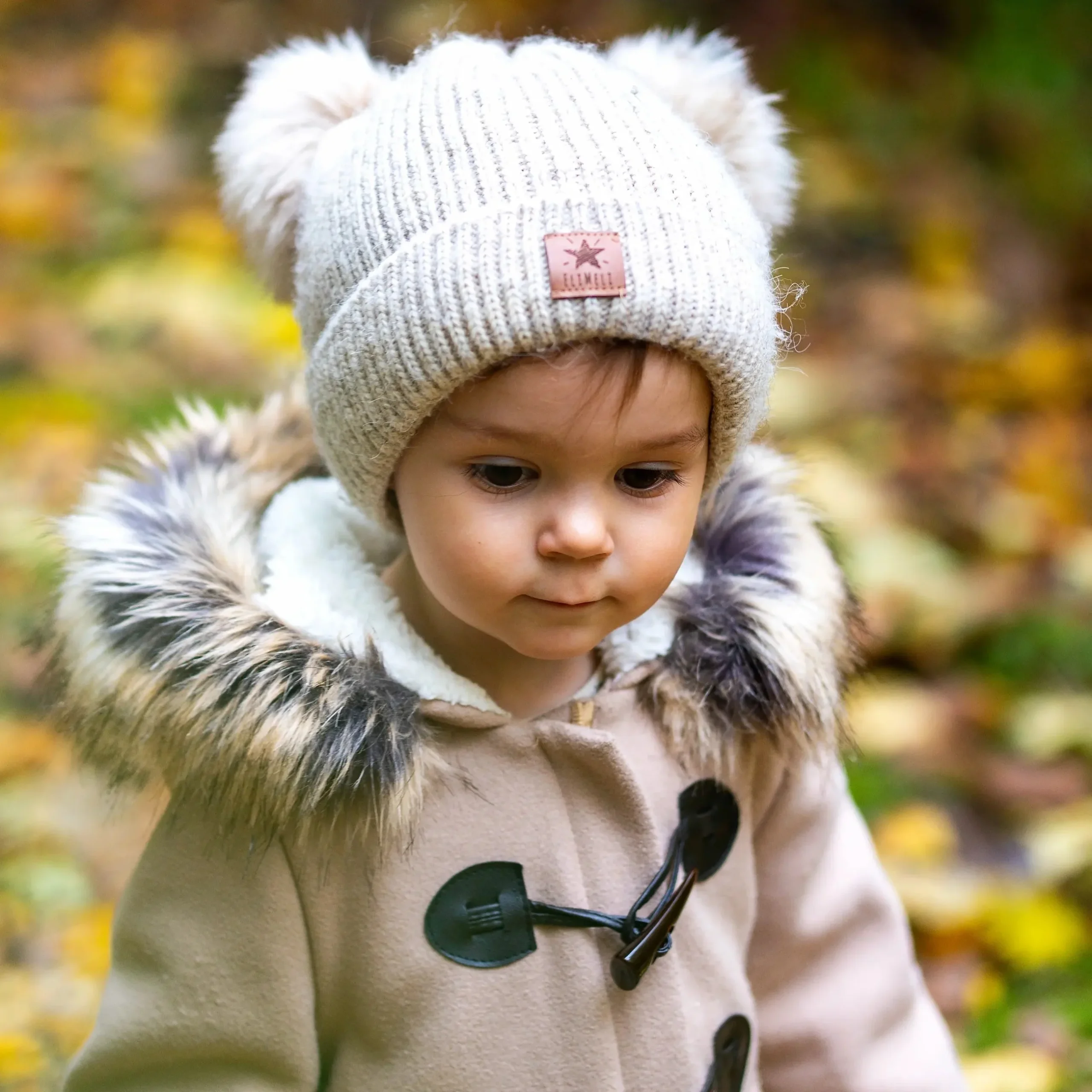 Kind mit beiger Mütze und Felljacke steht im herbstlichen Wald und blickt nach unten.