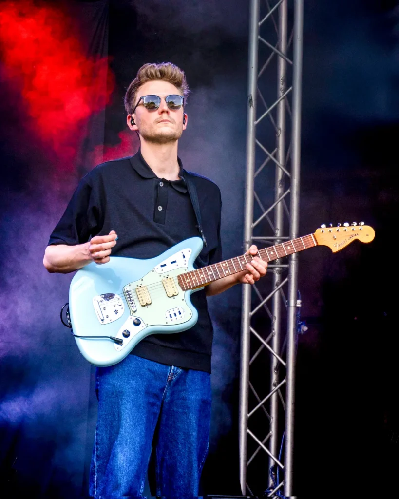Gitarrist mit Sonnenbrille spielt hellblaue E-Gitarre auf einer Bühne im bunten Licht.