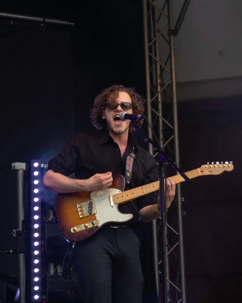 Gitarrist mit Sonnenbrille singt und spielt E-Gitarre auf einer Bühne.