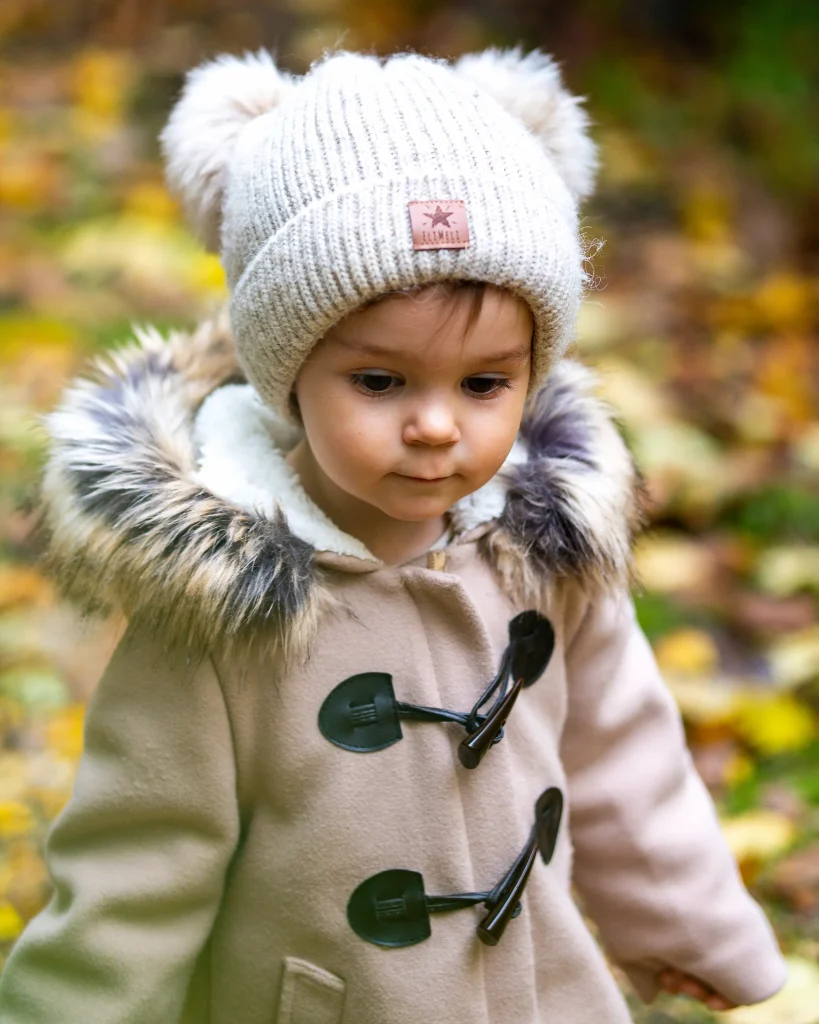 Kind mit beiger Mütze und Felljacke steht im herbstlichen Wald und blickt nach unten.