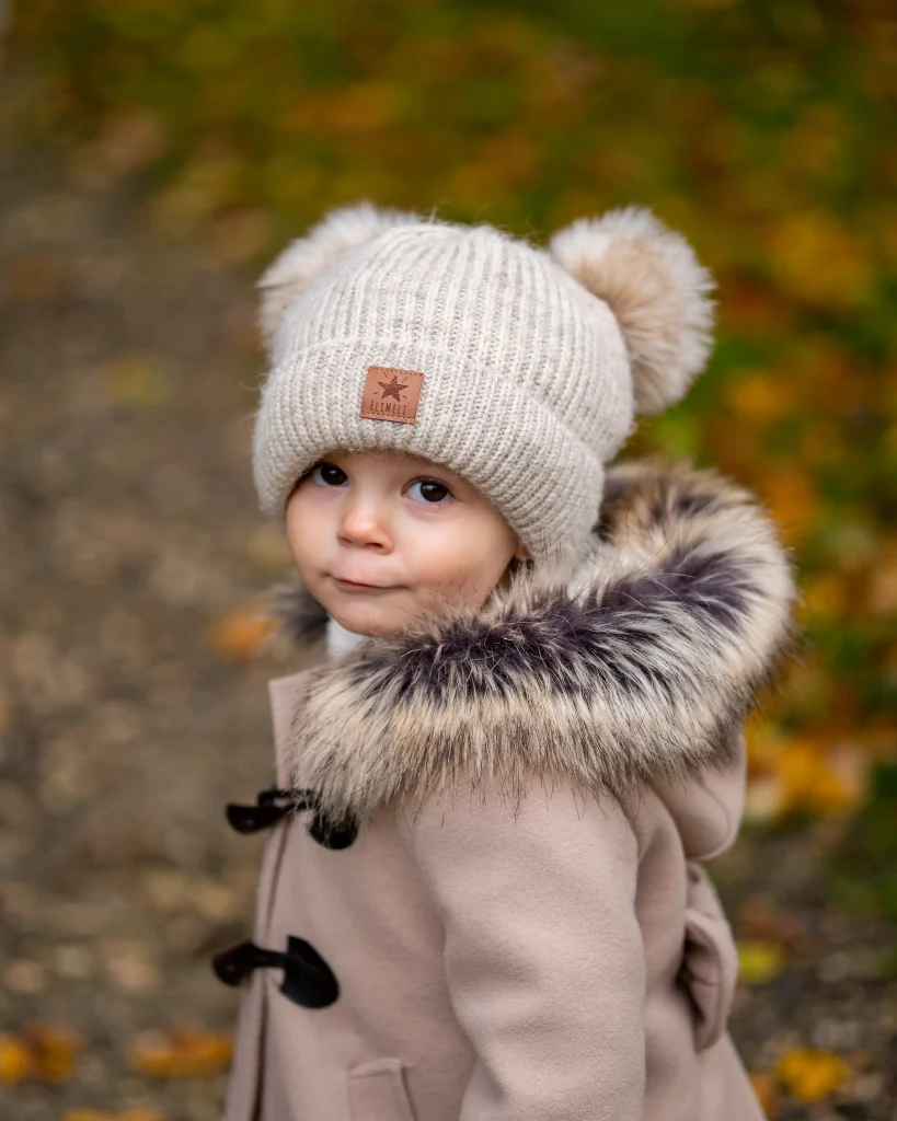 Kind mit beiger Mütze und Felljacke blickt in die Kamera, im herbstlichen Wald.
